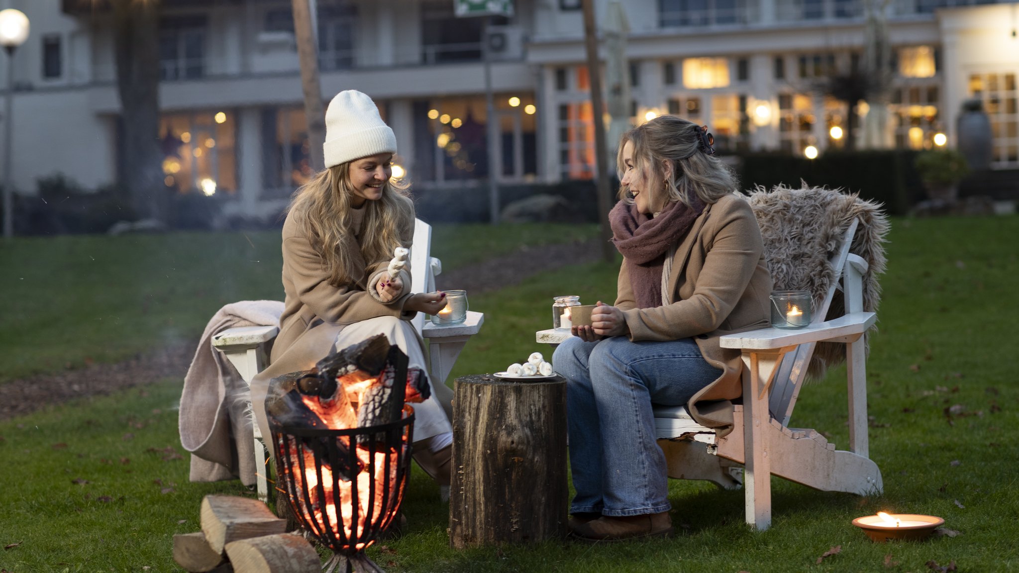 Twee dames roosteren marshmallows bij een kampvuur in de sneeuw, genietend van de hygge-sfeer bij Hotel de Bilderberg in de winter.