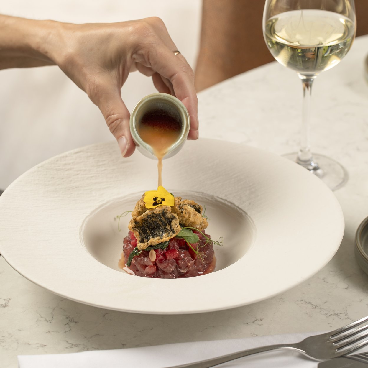 Hand giet gouden saus over tartaar met gegrilde vis, geserveerd met witte wijn in elegant restaurant.