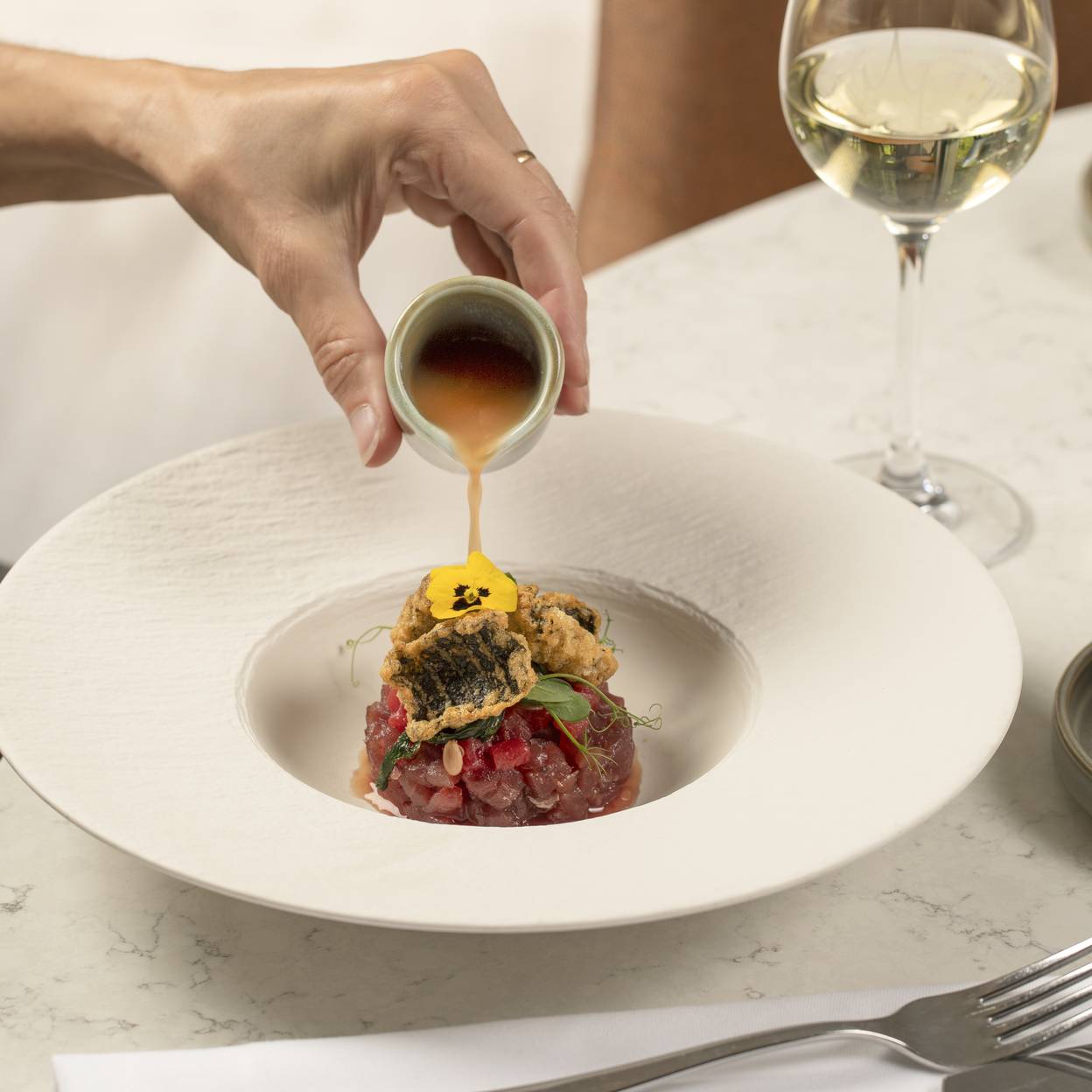 Hand giet gouden saus over tartaar met gegrilde vis, geserveerd met witte wijn in elegant restaurant.