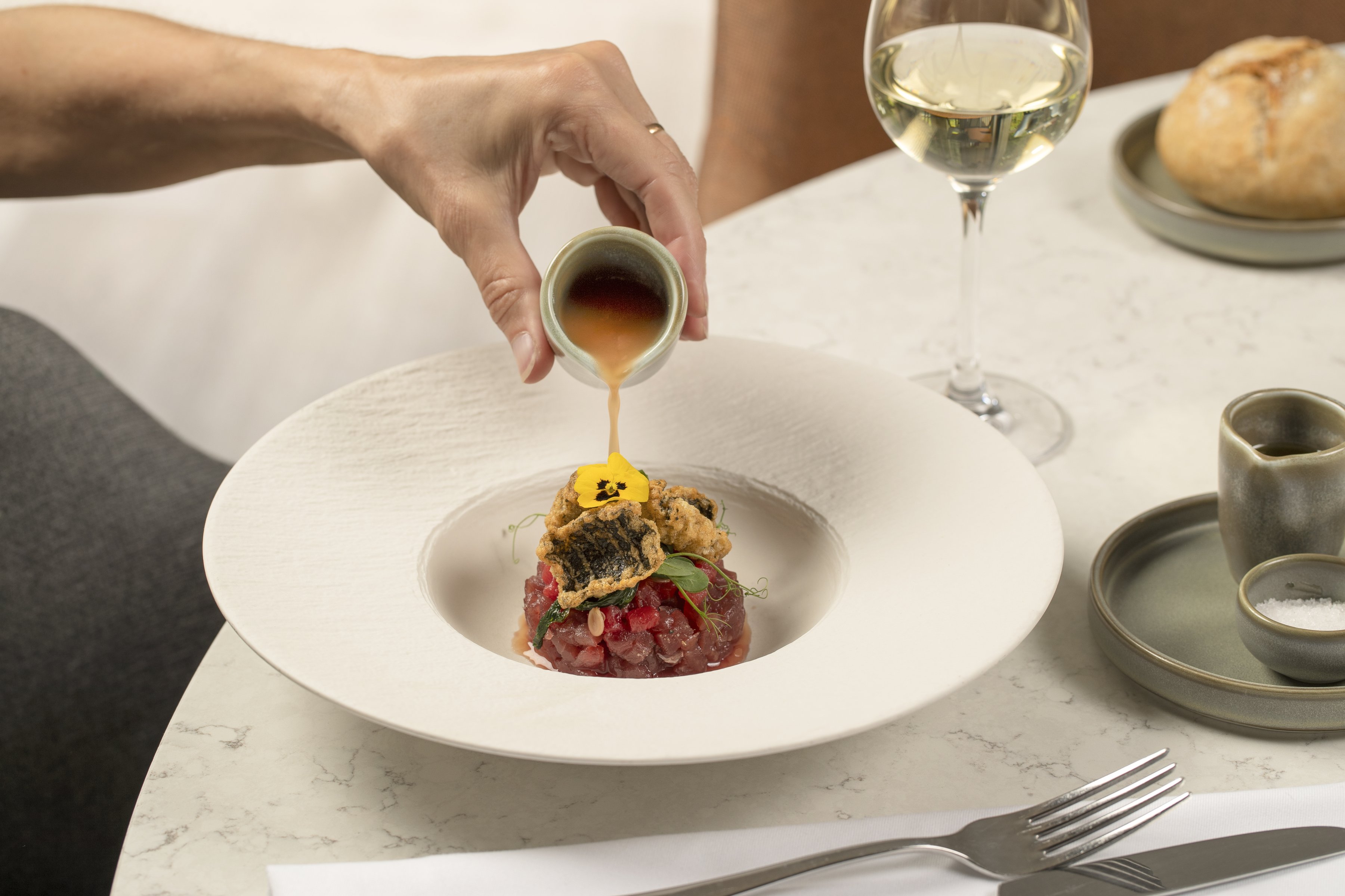 Hand gießt goldene Sauce über Tartar mit gegrilltem Fisch, serviert mit Weißwein in elegantem Restaurant.