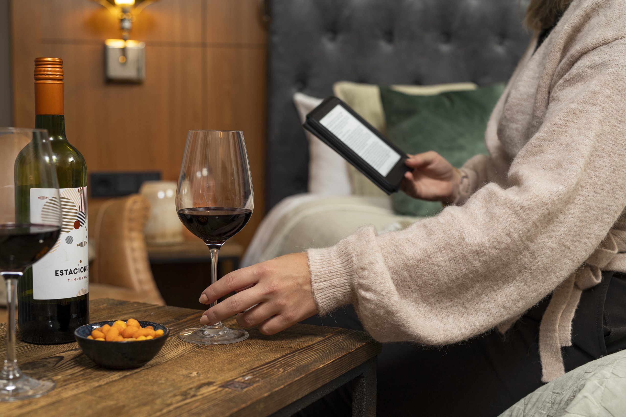 Een vrouw in een hotelkamer dat aan het lezen is op haar e-reader en ze pakt de een glas rode wijn.